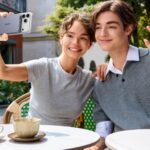 A smiling young man and woman sitting at an outdoor cafe, with the woman taking a selfie using an iPhone attached to a UGREEN magnetic power bank while the man waves at the camera