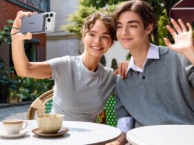 A smiling young man and woman sitting at an outdoor cafe, with the woman taking a selfie using an iPhone attached to a UGREEN magnetic power bank while the man waves at the camera