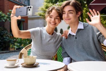 A smiling young man and woman sitting at an outdoor cafe, with the woman taking a selfie using an iPhone attached to a UGREEN magnetic power bank while the man waves at the camera
