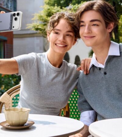 A smiling young man and woman sitting at an outdoor cafe, with the woman taking a selfie using an iPhone attached to a UGREEN magnetic power bank while the man waves at the camera