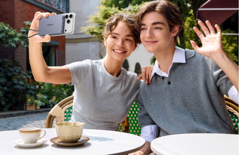 A smiling young man and woman sitting at an outdoor cafe, with the woman taking a selfie using an iPhone attached to a UGREEN magnetic power bank while the man waves at the camera